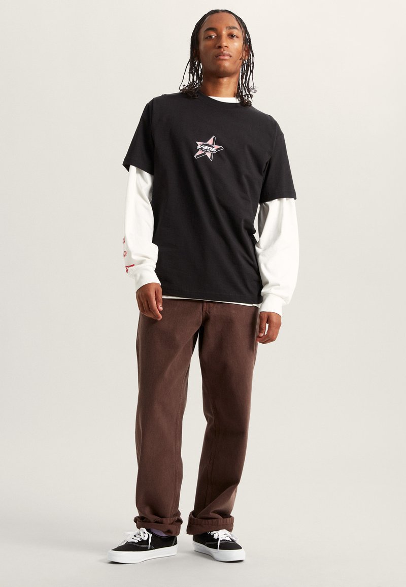 Czarny t-shirt z graficznym wzorem w gwiazdę i logo "Vans", nałożony na biały T-shirt z długim rękawem. Czekoladowe szerokie spodnie i czarne sneakersy z białą podeszwą.