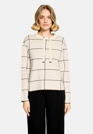 Femme portant un pull crème avec des lignes foncées en quadrillage et un détail noué à l'avant, associé à un pantalon noir, debout devant un fond uni.