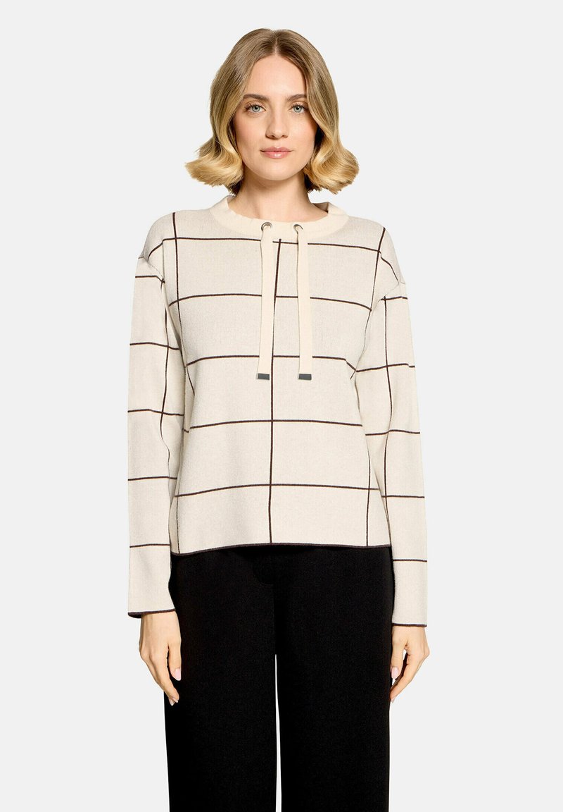 Femme portant un pull crème avec des lignes foncées en quadrillage et un détail noué à l'avant, associé à un pantalon noir, debout devant un fond uni.