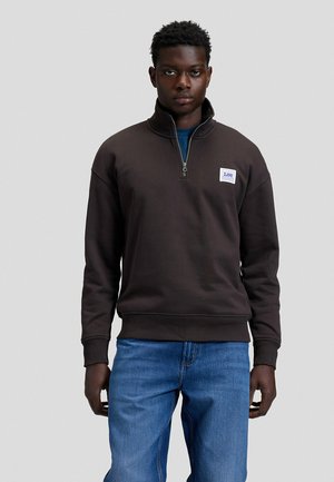 Man standing against plain background wearing a black quarter-zip sweatshirt with a Lee logo patch and blue jeans, facing forward.