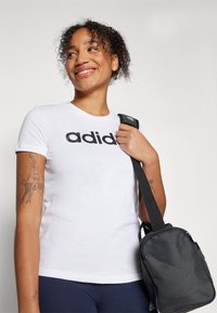 Bílý bavlněný tričko s výrazným černým logem "adidas", doplněné malou černou taškou s texturovaným povrchem a nastavitelným popruhem.