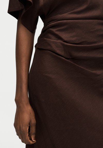 Άτομο με σκούρο δέρμα που φοράει σκούρο καφέ, κοντομάνικο φόρεμα με ρυτιδωμένο ύφασμα στη μέση, το χέρι χαλαρό δίπλα στο σώμα.