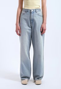 Jean large bleu clair en denim, avec une taille haute, des poches avant et un léger effet vintage. Porté avec un haut ajusté jaune.