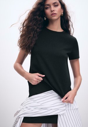 Schwarzes Baumwoll-T-Shirt mit kurzen Ärmeln, Rundhalsausschnitt und slim Fit. Getragen mit einem weißen gestreiften Stoff, der um die Taille drapiert ist und einen Kontrast in der Textur zeigt.