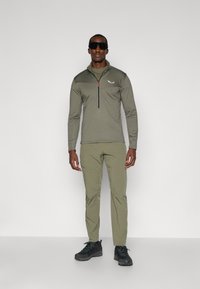 Olivegrünes, langärmliges Zip-Up-Oberteil mit hohem Kragen, kombiniert mit olivfarbenen Hosen. Ausgestattet mit schwarzen Wanderschuhen und dunklen Sonnenbrillen.