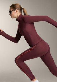 Bordo sportska odjeća s visokim ovratnikom, dugim rukavima i narančastim bočnim prugama. Izrađena od rastezljivog materijala, dizajnirana za pokret.