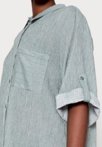 Camicia button-up a maniche corte di colore verde chiaro con righe verticali. Presenta una tasca sul petto e maniche arrotolate con dettagli a bottoni.