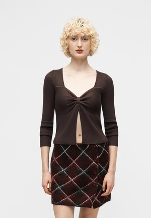 Ξανθιά γυναίκα με σγουρά μαλλιά που φοράει καφέ ριμπ μπλουζάκι με στρίφωμα μπροστά και κόκκινη-μαύρη καρό φούστα, μπροστά σε λευκό φόντο.