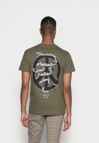 Camiseta verde oliva con un diseño en blanco y negro de una serpiente que rodea un símbolo de paz en la parte posterior. Mangas cortas, cuello redondo, fabricada en algodón.
