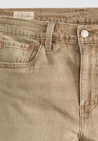 Jeans de ganga bege com um acabamento texturizado. Apresenta um pequeno bolso frontal, rebites de cobre e uma etiqueta da marca na cinta.