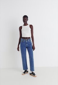 Balta bezmaku kultūra, zili augstās jostas jeans un melnas kedas ar baltajiem akcentiem. Modelis stāv pret vienkāršu gaišu fonu.