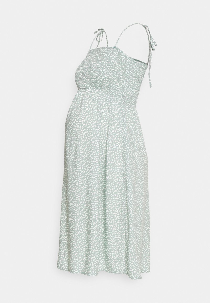 Robe de maternité vert clair avec imprimé de feuilles blanches, fines bretelles et corsage smocké, s'évasant en une jupe jusqu'au genou.