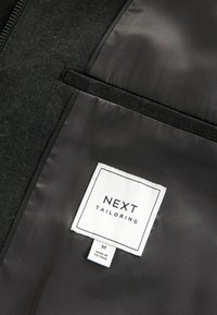 Czarna, gładka podszewka z kieszenią na zamek. Metka pokazuje "NEXT TAILORING", rozmiar M oraz "WYPRODUKOWANE W WIETNAMIE."