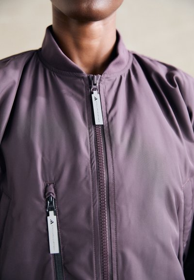 Lila bomberdzseki, elöl cipzárral, bordás gallérral és oldalsó zsebekkel, márkázott cipzárhúzóval. Simább, könnyű anyag.