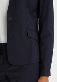 Námorně modrý přizpůsobený blazer se dvěma knoflíky, jednou náprsní kapsou a hladkou texturou, součást dvoudílného obleku.