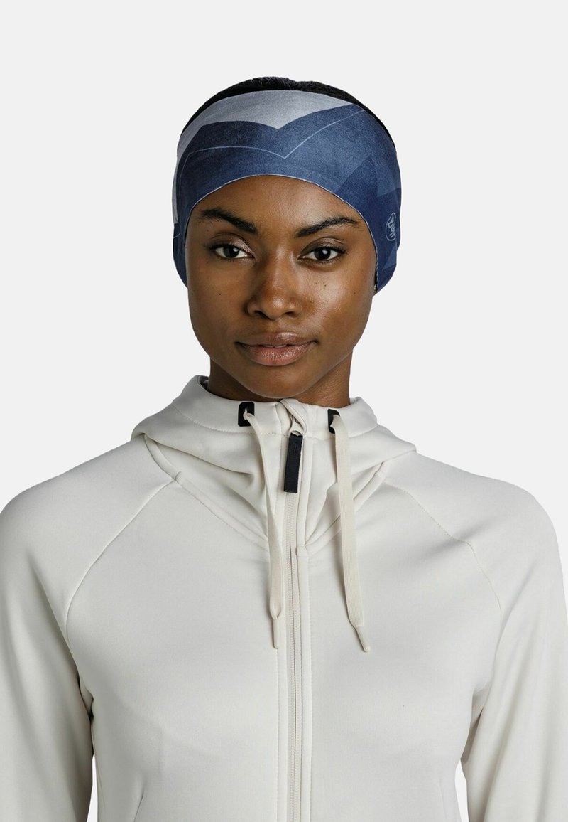 Bandeau dans des tons de bleu avec des motifs géométriques ; fabriqué en tissu doux et extensible. Porté par une personne dans une veste zippée couleur crème.