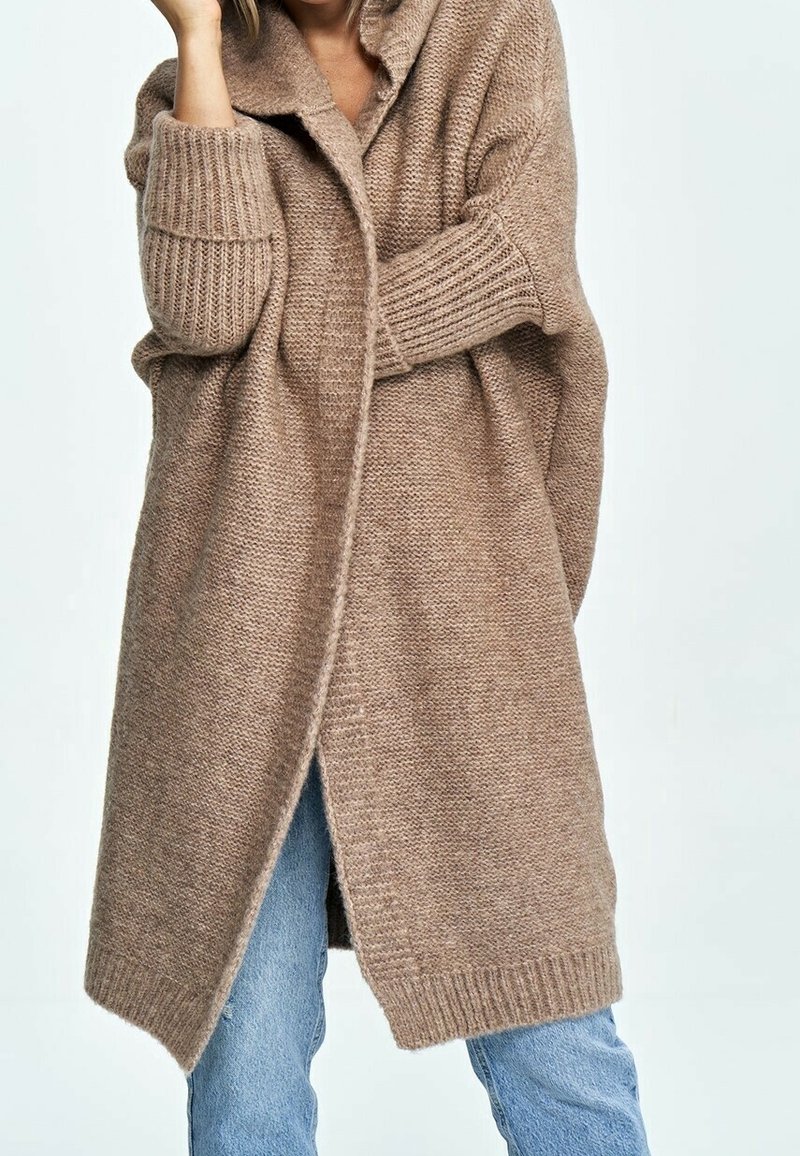 Beige long cardigan made of textured knitted material, featuring a collar, front button closure, and ribbed cuffs, layered over blue jeans.