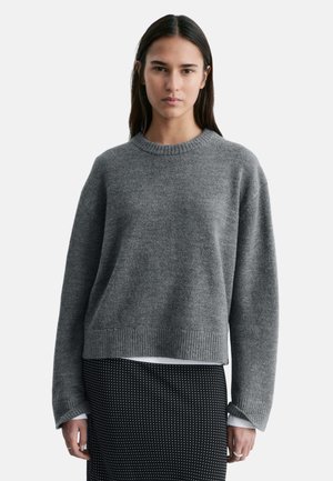 Ung kvinde med langt mørkt hår iført en grå strikket sweater over en hvid skjorte og et sort nederdel med hvide polka prikker, stående fremad.