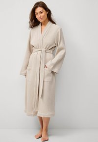 Beige badjas met wafelstructuur, lange mouwen, een ceintuur en twee voorzakken. Korte lengte tot de knieën. Zacht, lichtgewicht materiaal.