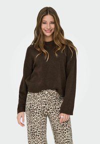 Junge Frau lächelt, trägt einen dunkelbraunen Pullover und eine Hose mit Leopardenmuster, hat lange, wellige Haare und einen Ring an ihrer rechten Hand.