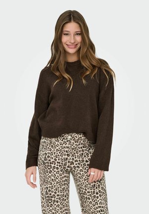 Mlada žena se smiješi, nosi tamnosmeđu vestu i hlače sa leopard printom, ima dugu valovitu kosu i prsten na desnoj ruci.