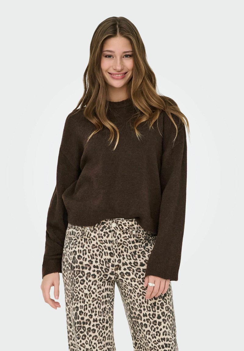 Junge Frau lächelt, trägt einen dunkelbraunen Pullover und eine Hose mit Leopardenmuster, hat lange, wellige Haare und einen Ring an ihrer rechten Hand.
