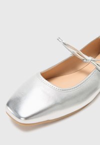 Silver metallic ballet flat with a square toe, smooth texture, and a thin ankle strap tied with a bow. Light tan insole visible.