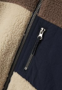 Tessuto navy trapuntato con pannelli in pile marrone e crema; presenta una tasca con zip e hardware in argento per una conservazione sicura.