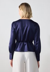 Marine satengbluse med pufferærmer, rynket talje og knappebånd; glatt tekstur og enkelt design. Brukt med lyse bukser.