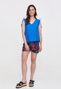 Niebieski bezrękawnik z dekoltem w kształcie V i wiązaniami, w połączeniu z wielokolorowymi szortami w kwiaty oraz czarnymi sandałami na koturnie z zapięciem.
