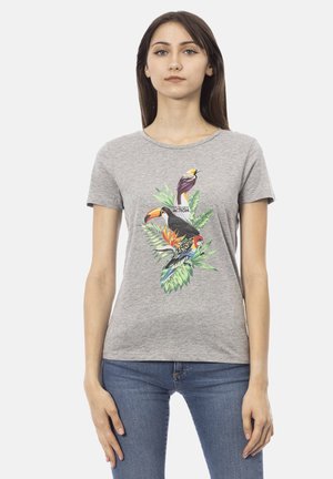 Jonge vrouw draagt een grijs T-shirt met een kleurrijke print van tropische vogels en groene bladeren, gecombineerd met een blauwe spijkerbroek, tegen een effen achtergrond.