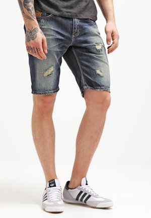Homme portant un short en jean bleu déchiré, un t-shirt noir et des baskets blanches Adidas à rayures noires, debout avec la main gauche tatouée visible.
