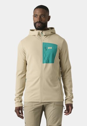 Man draagt een beige zip-up capuchonvest met een teal borstzak en bijpassende beige broek, staand tegen een effen achtergrond.