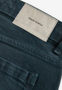 Donkerblauwe denimstof met een gestikt label met de tekst "DENIM SUPPLY." Heeft een achterzak met zichtbare stiksels.