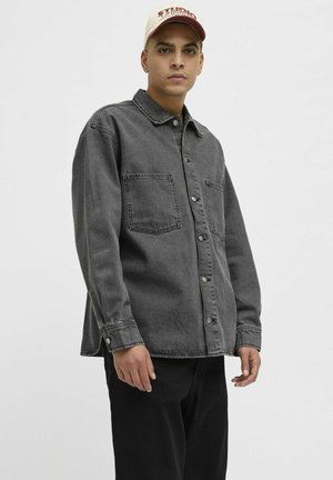 Gray denim shirt with long sleeves, traditional collar, two front pockets, and snap-button closure, paired with black pants and a cream baseball cap.