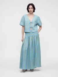 Femme debout portant un haut à manches courtes rayé bleu et vert avec des liens devant et une jupe longue, plissée, assortie avec des chaussures blanches.
