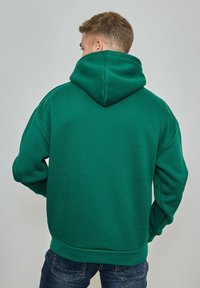Personne portant un sweat à capuche vert et un jean bleu, vue de dos sur un fond gris uni.