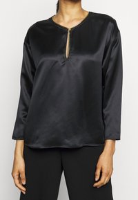Blouse longue en satin noir à manches longues avec un col en trou de serrure orné de détails dorés et une texture lisse et brillante. Design simple, coupe décontractée.
