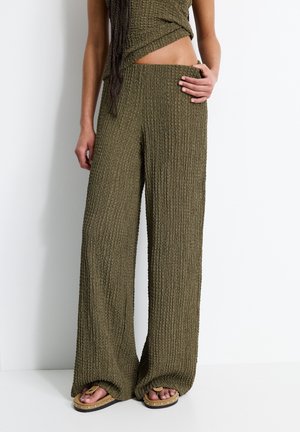 Woman wears olive green textured wide-leg pants and matching top, showing bare midriff, with tan studded sandals on a white background.