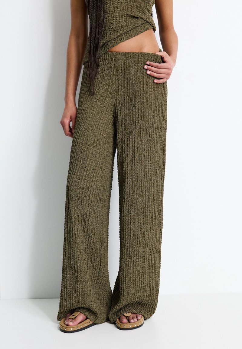 Femme porte un pantalon large texturé vert olive et un haut assorti dévoilant le ventre, avec des sandales cloutées couleur caramel sur fond blanc.