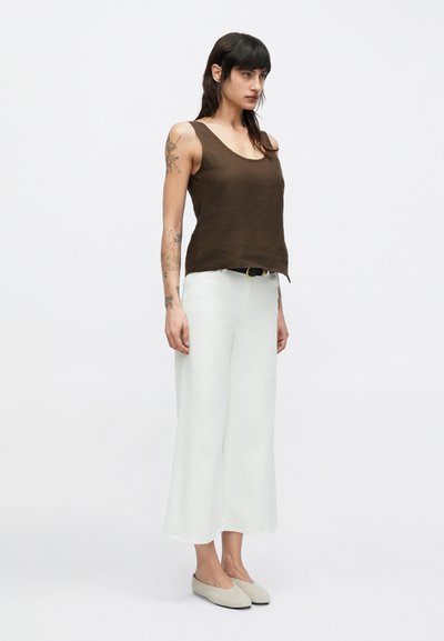 Καφέ λινό φανελάκι χωρίς μανίκια, λευκό παντελόνι με φαρδιά μπατζάκια και ανοιχτό γκρι παπούτσια χωρίς κορδόνια. Ορατά τατουάζ στο αριστερό μπράτσο· μινιμαλιστικός σχεδιασμός και ουδέτερα χρώματα.