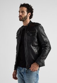 Zwarte leren jas met rits aan de voorkant, twee voorzakken en drukknoopdetails op de schouders. Gladde textuur, getailleerd design, gecombineerd met blauwe jeans.