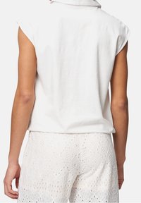 Haut sans manches en coton blanc avec un col rond, porté avec un short blanc à motifs orné de découpes complexes.