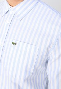Chemise rayée verticale bleu clair et blanc avec une poche avant et un petit logo crocodile vert sur la poche.