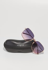 Ochelari de soare cu accente aurii și lentile violet cu degradé, odihnindu-se pe un toc oval negru cu fermoar, având inscripționat numele mărcii "vogue."