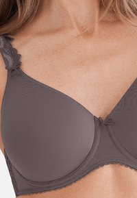 Sottocosto marrone senza cuciture con spalline lavorate in pizzo, coppe sagomate e un sottile fiocco centrale. Texture liscia con finitura opaca.