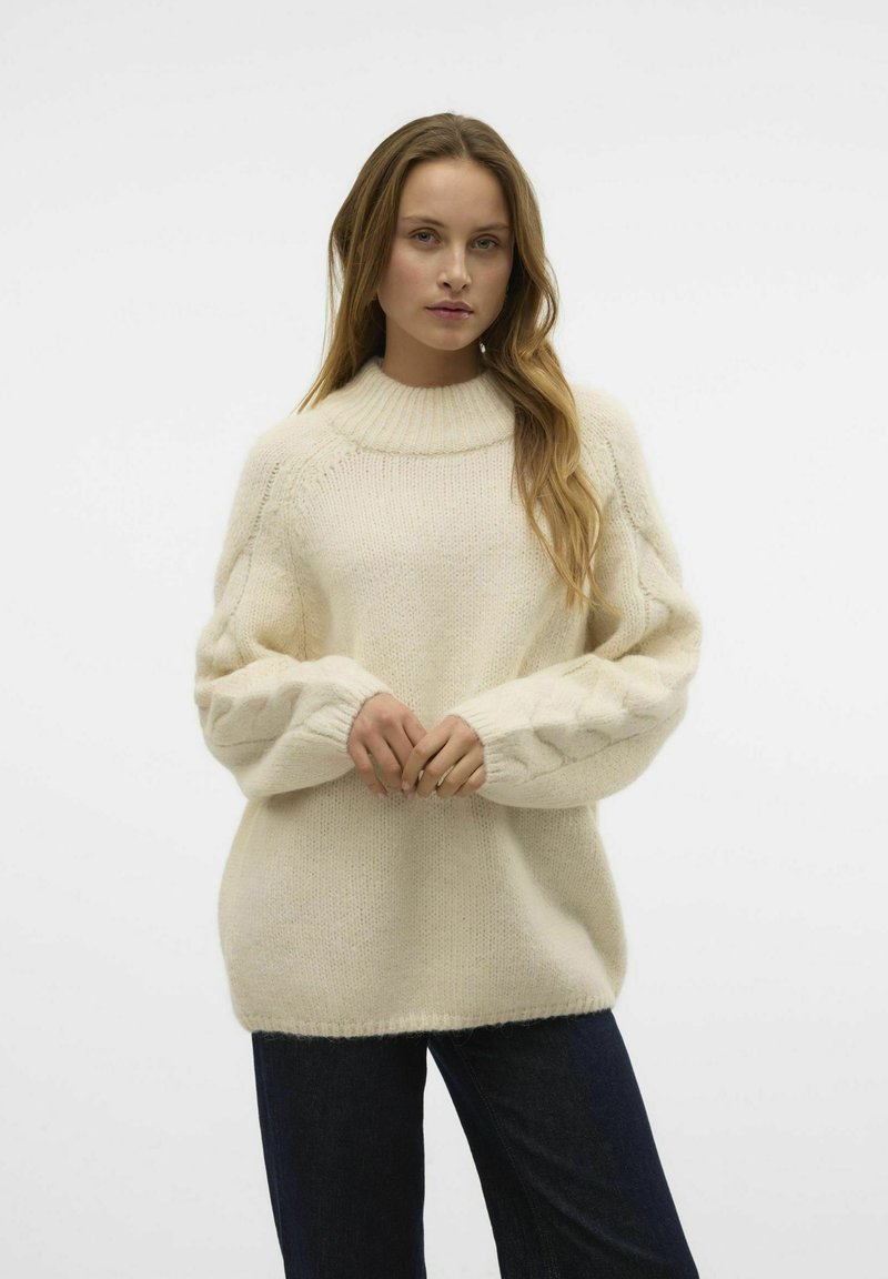 Cremiger Strickpullover mit hohem Rippkragen, strukturierten Zopfmusterärmeln und lockerer Passform. Kombiniert mit dunklen Denim-Hosen.