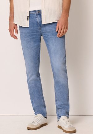 Homme portant un jean slim bleu clair, des baskets blanches et une chemise beige, debout devant un fond uni.
