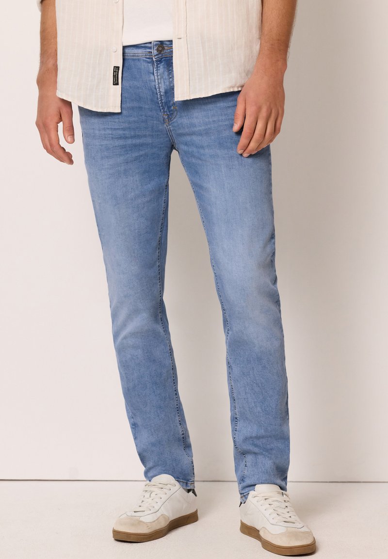 Homme portant un jean slim bleu clair, des baskets blanches et une chemise beige, debout devant un fond uni.