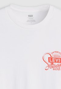 Balta medvilnės marškinėliai su ryškiu oranžiniu Levi's logotipu, tekstu ir dizaino akcentais šalia apykaklės, trumpomis rankovėmis ir standartu tinka.